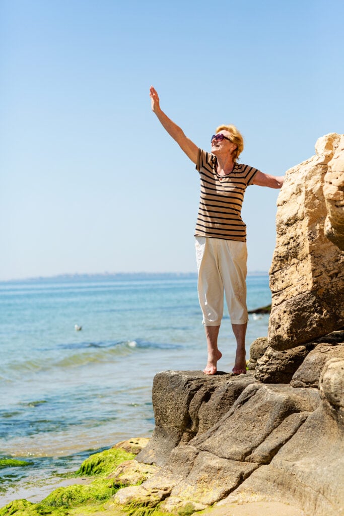 Elderly Attractive Smiling Woman Standing On A Rock On The Seashore